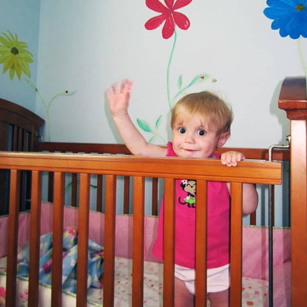 toddler waving from crib with flowers painted on the wall in background